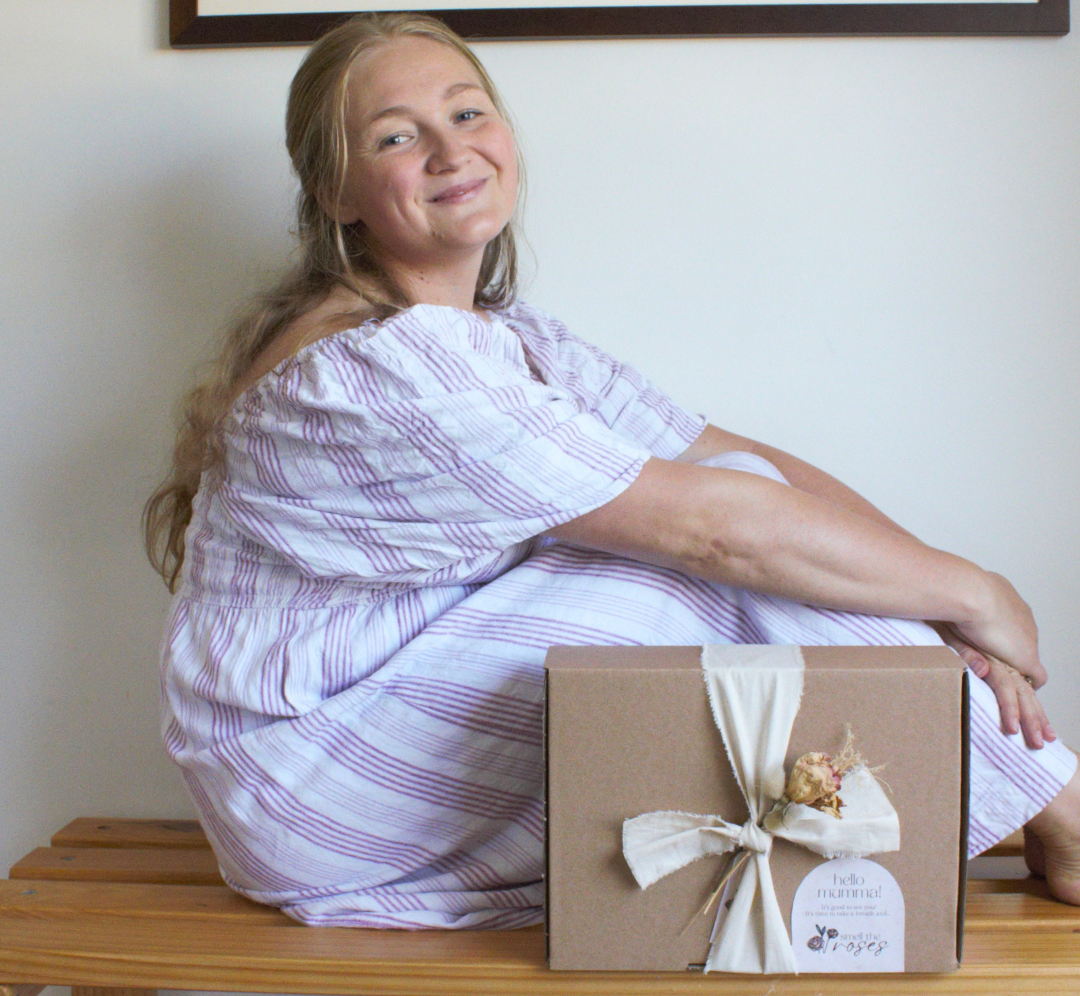 A woman sitting on a wooden bench wearing a white and purple striped dress, with a brown gift box tied with a cream ribbon placed in front of her. A framed landscape print hangs on the wall behind her.