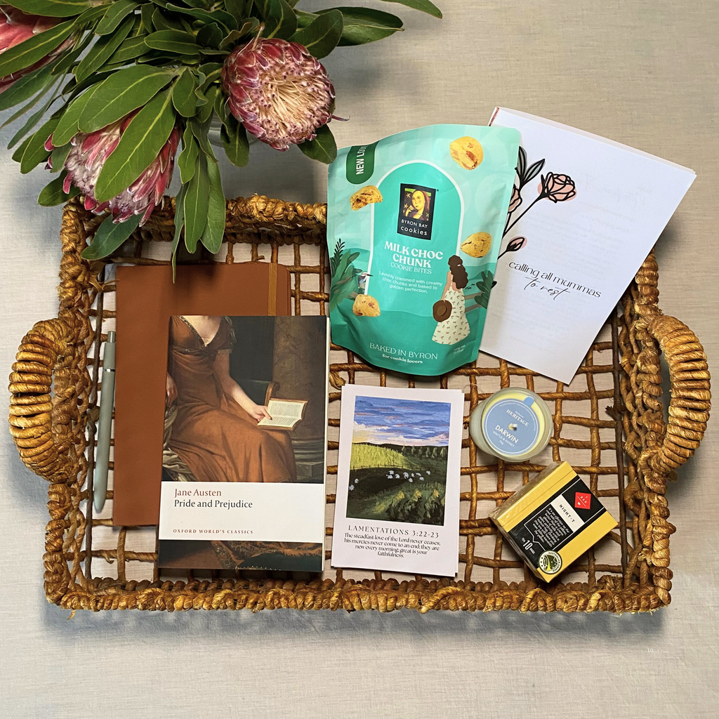 A curated gift tray arrangement on a woven wicker tray, featuring a teal pouch of milk chocolate biscuits, a white card with landscape art, a pride and prejudice book, a small candle, a notebook, and a box of night T from the T bar, styled with green leaves and pink flowers on a light fabric background.