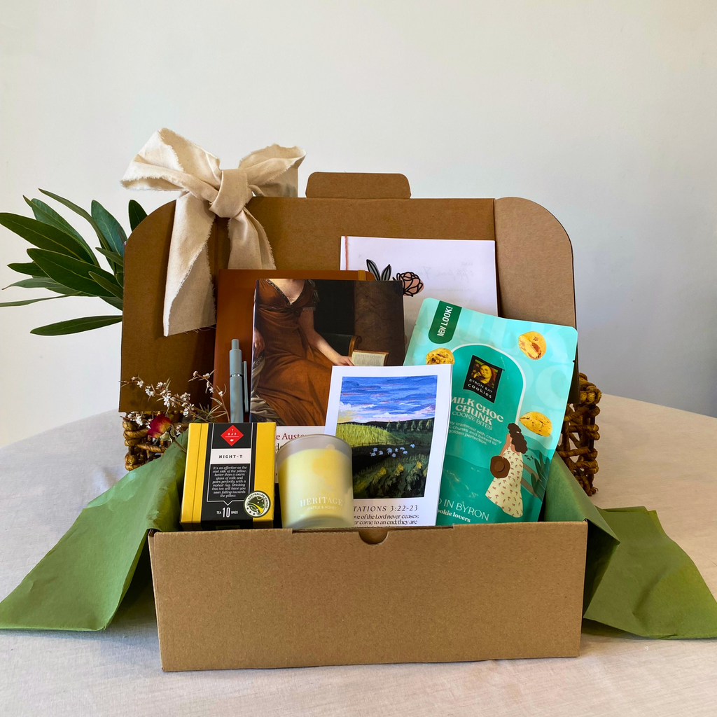 A curated gift box set in a brown cardboard box with a cream ribbon, filled with a green tea bag, a candle, a small picture frame, and a postcard, displayed on a light-colored table.