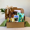 A curated gift box set in a brown cardboard box with a cream ribbon, filled with a green tea bag, a candle, a small picture frame, and a postcard, displayed on a light-colored table.