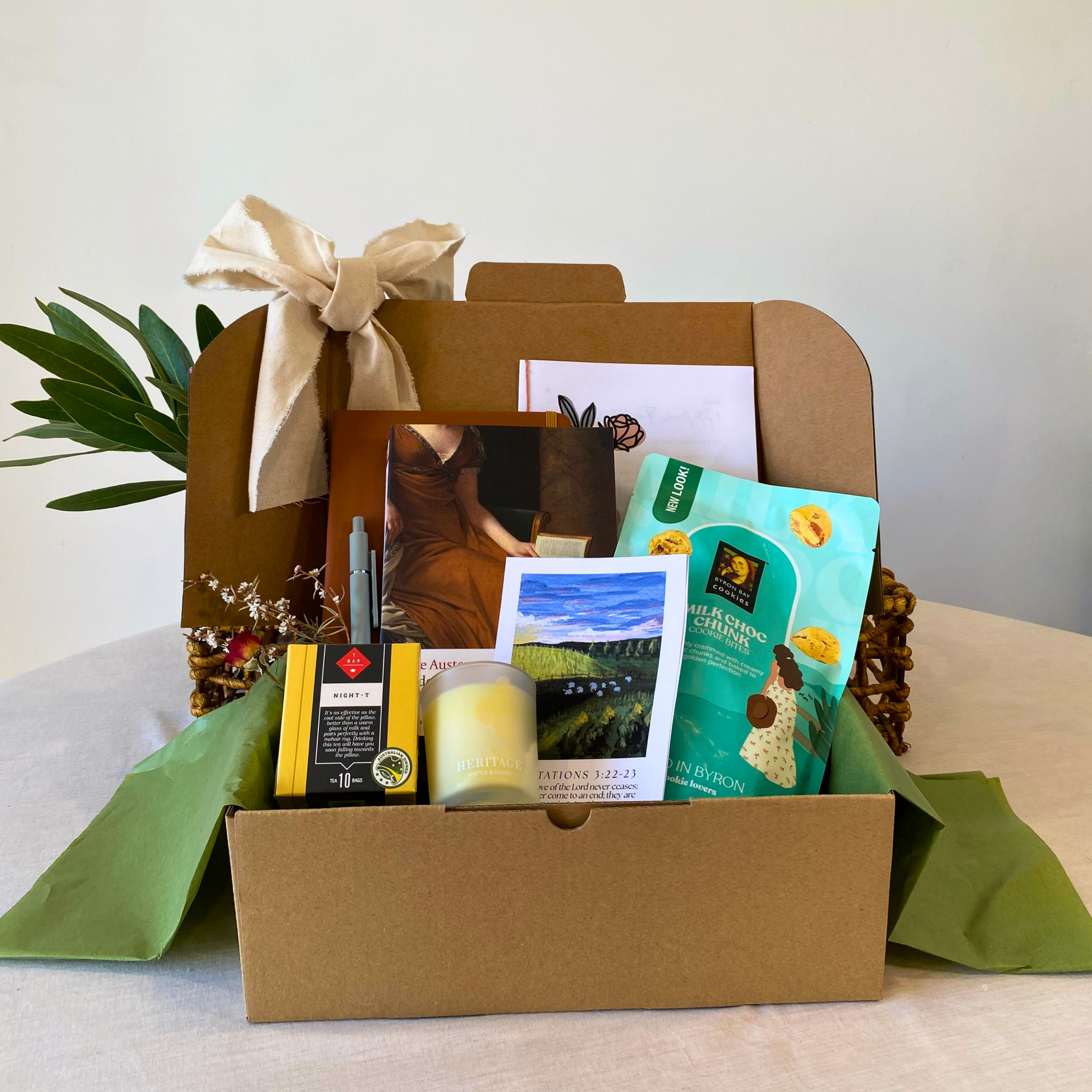 A curated gift box set in a brown cardboard box with a cream ribbon, filled with a green tea bag, a candle, a small picture frame, and a postcard, displayed on a light-colored table.