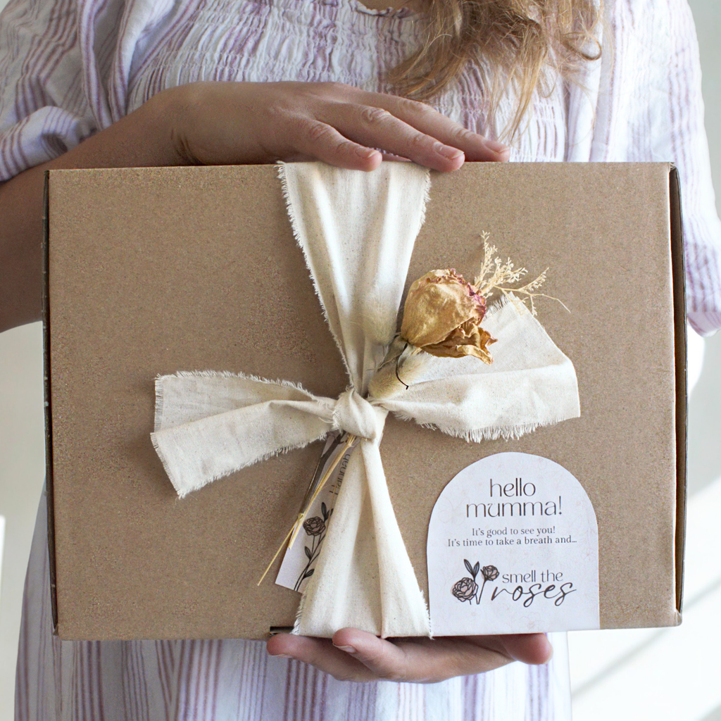 Brown gift box with a off white ribbon tied around the box with a bouquet of dried flowers, held by a woman wearing a striped dress pink an linen white dress.