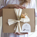 Brown gift box with a off white ribbon tied around the box with a bouquet of dried flowers, held by a woman wearing a striped dress pink an linen white dress.